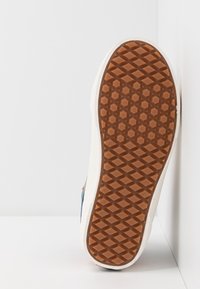 Semelle extérieure en caoutchouc avec un motif hexagonal texturé, bord blanc crème et dessus en tissu coloré. Vue latérale sur un fond uni.