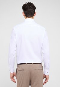 Weißes Langarm-Hemd mit Button-Down-Kragen und Manschetten Details, kombiniert mit hellbraunen Maßhosen. Glatte Stofftextur.
