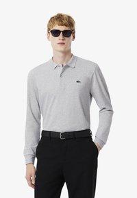 Grå långärmad polo skjorta med krage, med en liten marinblå logotyp, i kombination med svarta byxor och ett svart bälte. Svarta solglasögon på.