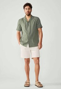 Camicetta a maniche corte, con bottoni, di colore verde oliva con strisce verticali bianche, abbinata a pantaloni corti bianchi e sandali beige. Texture morbida, design casual.