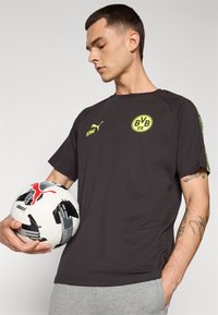 Sort kortærmet t-shirt med et gult logo og detaljer, holdt ved siden af en hvid fodbold med sorte og røde grafikker, der viser et sporty design.