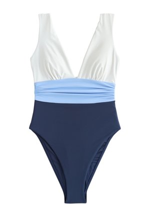 Femeie într-un costum de baie în blocuri de culoare, ținând un prosop în dungi, stând pe un doc de lemn cu bărci cu pânze și apă în fundal.