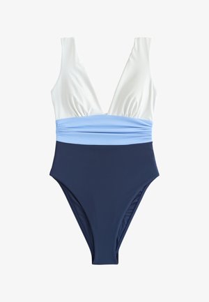 Femme en maillot de bain color-block tenant une serviette rayée, debout sur un quai en bois avec des voiliers et de l'eau en arrière-plan.