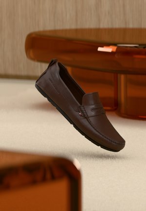 Pantof maro din piele, tip loafer, plutind deasupra covorului bej, cu mobilier modern din sticlă cu nuanță chihlimbar în fundal.