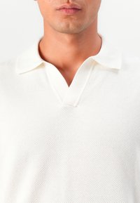 Weißes Poloshirt mit einem strukturierten Strickmuster, kurzen Ärmeln und tiefem V-Ausschnitt. Verfügt über einen umgeschlagenen Kragen und einen glatten Stoff.