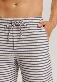 Gestreifte Badeshorts in Beige und Marineblau, aus strukturiertem Stoff gefertigt. Verfügt über einen Kordelzugbund und eine Seiten Tasche.