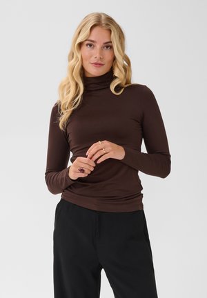 Femme aux longs cheveux blonds portant un col roulé marron et un pantalon noir, debout les mains près de la taille devant un fond uni.