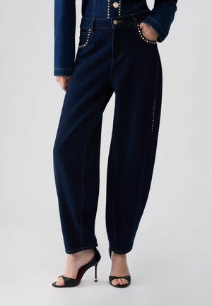 Jean bleu foncé à jambes larges avec détails cloutés sur les poches et les côtés, porté avec des talons hauts ouverts noirs et un top en denim assorti.