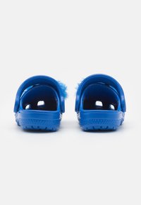 Pár modrých gumových dřeváků s huňatým modrým podšívkou, zobrazený zádní stranou na bílé ploše.