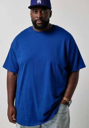 Mann mit Bart, der eine blaue Los Angeles Baseballkappe, ein blaues T-Shirt, helle Jeans und eine Armbanduhr trägt, steht vor einem einfarbigen Hintergrund.