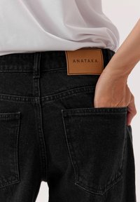 Czarne dżinsy z denimu z skórzaną metką z napisem "ANATAKA", posiadające dwie tylne kieszenie oraz zbliżenie na pas.