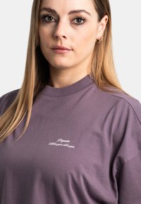 Camiseta de algodón morada con cuello redondo y mangas cortas. Presenta texto bordado en blanco en la parte delantera y una textura suave y lisa.