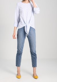 Hellbluse mit weißen Streifen und vornem Knoten, kombiniert mit engen blauen Jeans und gelben Ballerinas. Lässiges Outfit mit lockerem Schnitt.