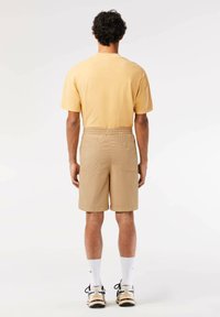 Pantalones cortos beige con una cinturilla elástica, dos bolsillos traseros, combinados con una camiseta de color amarillo claro, usados con calcetines blancos y zapatos negros.