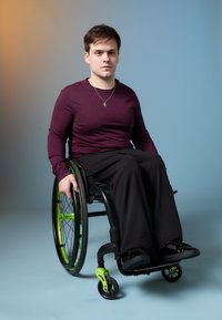Άτομο σε αναπηρικό καροτσάκι με πράσινους και μαύρους τροχούς, φορώντας μωβ μακρυμάνικη μπλούζα και μαύρα παντελόνια, καθισμένο σε έναν φθίνοντα φόντο.