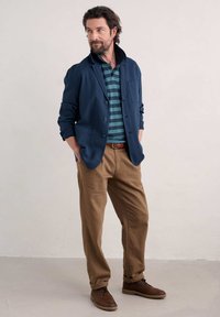 Blazer léger navy par-dessus un polo rayé bleu-vert et noir, associé à un pantalon beige et des chaussures marron à lacets. Design simple et moderne.