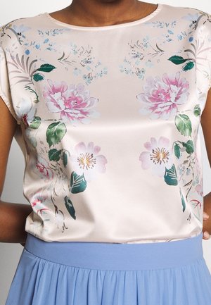 Frau trägt eine satinierte Bluse mit pinken und weißen Blumen, kombiniert mit einem hoch taillierten hellblauen plissierten Rock.