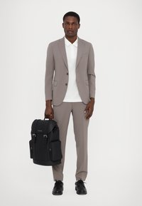 Costum gri deschis cu un sacou ajustat și pantaloni tapered, purtat peste o cămașă albă, asortat cu un rucsac din piele neagră cu accesorii din argint.