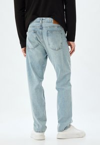 Jeans de denim azul claro con un corte relajado, bolsillos traseros visibles y un parche de cuero marrón en la cinturilla. Llevados con zapatos blancos.