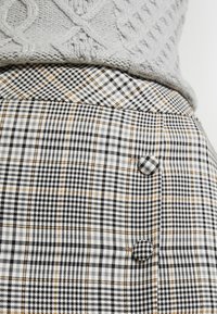 Un pull en tricot gris au-dessus d'une jupe à carreaux présentant des motifs noirs, blancs et orange, avec deux boutons ronds proéminents sur le côté.