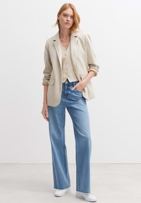 Blazer beige in lino con maniche arrotolate, abbinato a un gilet beige chiaro con bottoni e jeans chiari a vita alta. Sneakers bianche.