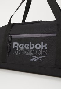 Bolsa de deporte negra con el logo gris de Reebok, con un cierre superior y un bolsillo con cremallera en el lateral. Fabricada con tela duradera y asas resistentes.