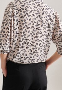 Ljusrosa blus med ett svart lövmönster, med lös passform och 3/4-långa ärmar. Matchad med svarta byxor som visar en avslappnad siluett.