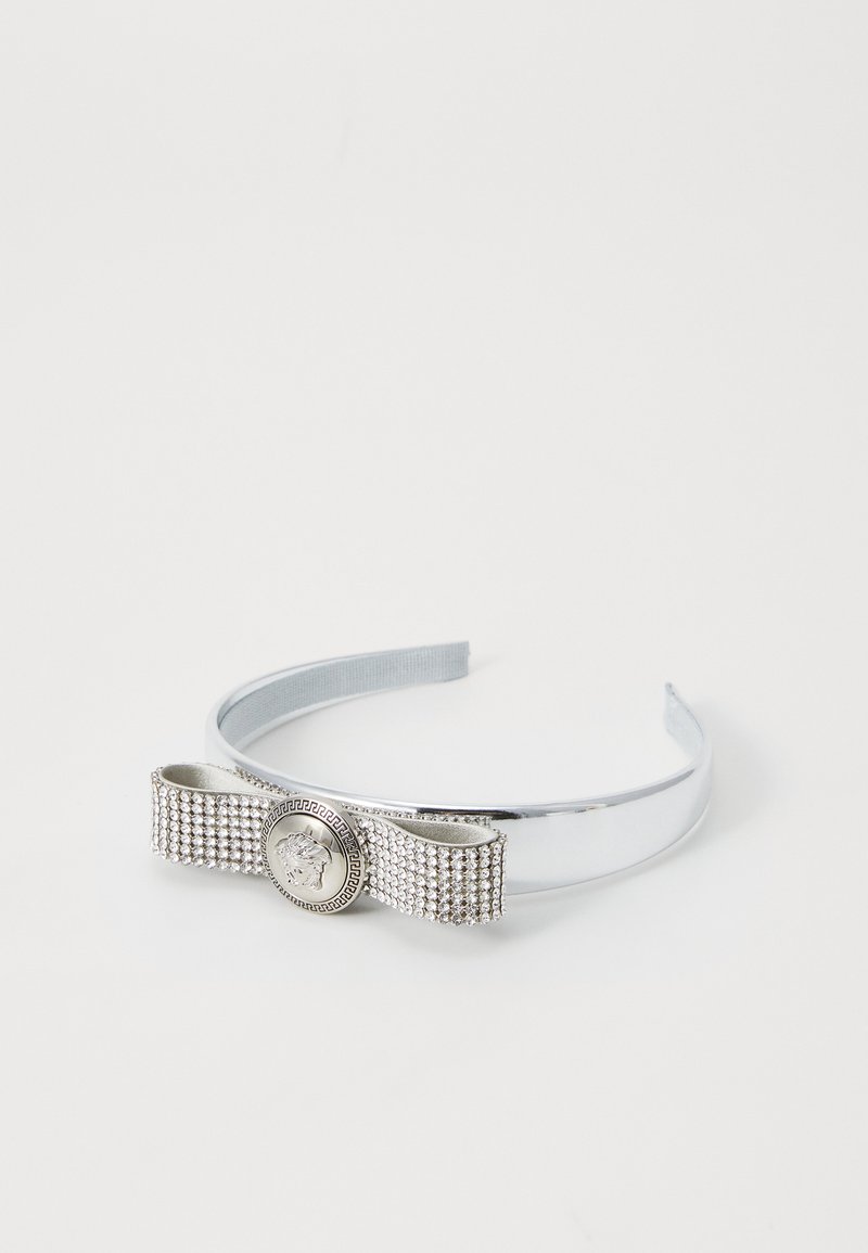 Sølv hårbånd med en sløjfe detalje, prydet med rhinsten og et rundt emblem i midten. Glat, reflekterende metaloverflade.