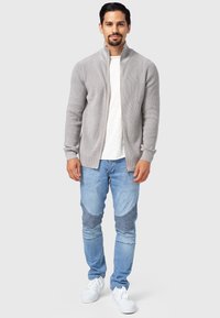 Suéter de cremallera acanalado gris claro sobre una camisa blanca, combinado con jeans azules desgastados con parches a rayas y zapatillas blancas.