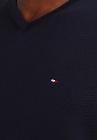 Pull en maille bleu marine à col en V, fabriqué en tissu texturé, avec un petit logo rouge, blanc et bleu sur le côté gauche de la poitrine.