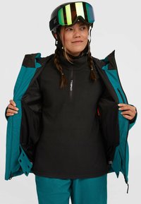 Tealgroene skijack met zwarte accenten, gedragen over een zwarte fleece laag. Voorzien van een rits aan de voorkant en verstelbare manchetten; inclusief skibril.