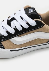 Zapatilla de ante en tonos marrón y negro con cordones blancos y logotipo, que presenta una franja lateral blanca y costuras negras sobre una suela de goma blanca.