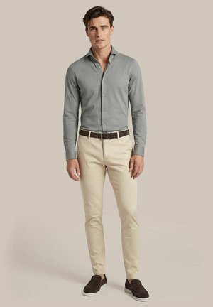 Man draagt een grijs overhemd met knopen, beige broek, bruine riem en donkere instapschoenen, staand tegen een effen beige achtergrond.