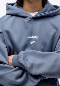 Sudadera gris con una gran impresión frontal que dice "STWD REC." Tejido texturizado con puños acanalados y un pequeño logotipo en la manga. Capucha ajustable.