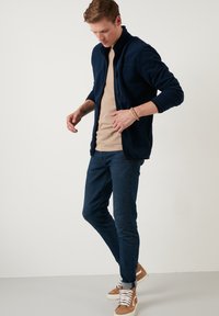 Navy fleece jacket with a high collar, front zippers, and textured finish, paired with tan t-shirt and dark blue slim-fit jeans.