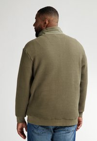 Olivgrön sweatshirt med textur och hög krage, ribbade ärmslut och nertill. Bärs över blå denimjeans och visar en avslappnad passform.