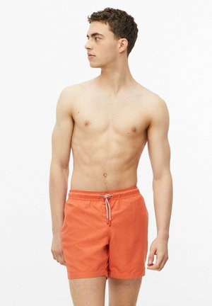 Jonge man staat shirtloos in oranje zwembroek met witte trekkoord, kijkt naar links tegen een effen achtergrond.