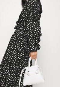 Musta mekko, jossa valkoinen pilkkukuosi, pitkät hihat ja kerätty vyötärö. Mekon kanssa pieni valkoinen tekonahkainen käsilaukku, jossa pyöreät kahvat.
