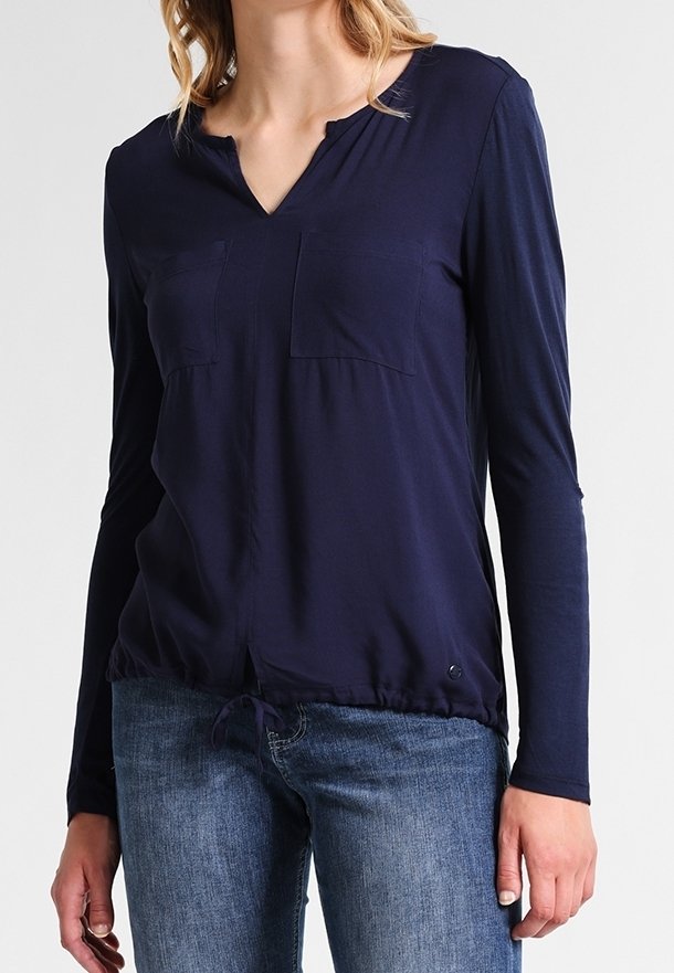 Top de manga larga azul marino con cuello en V, que cuenta con dos bolsillos en el pecho y un dobladillo con cordón. Tejido suave con un ajuste relajado. Llevado con vaqueros azules.
