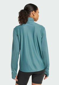 Long-sleeve athletic top in teal, featuring a high collar and textured fabric. Worn with black shorts, showing a relaxed fit and full sleeves.