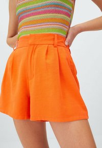 Shorts arancioni in cotone con design plissettato, vita alta e tasche laterali. Texture morbida e vestibilità sartoriale creano una silhouette rilassata.