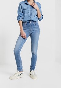 Jeans de mezclilla azul claro con corte ajustado, cintura alta y costuras en contraste. Combinados con una camisa de mezclilla azul claro y zapatillas blancas.