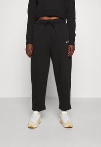 Svarta sweatpants med elastisk midja, sidofickor och en diskret vit Nike-logo på vänster lår, ihop med ljusa sneakers.