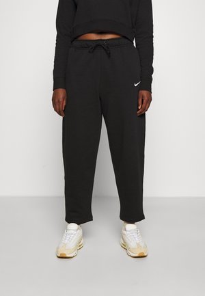 Black sweatpants with elastic waistband, side pockets, and a subtle white Nike logo on the left thigh, paired with light sneakers.
