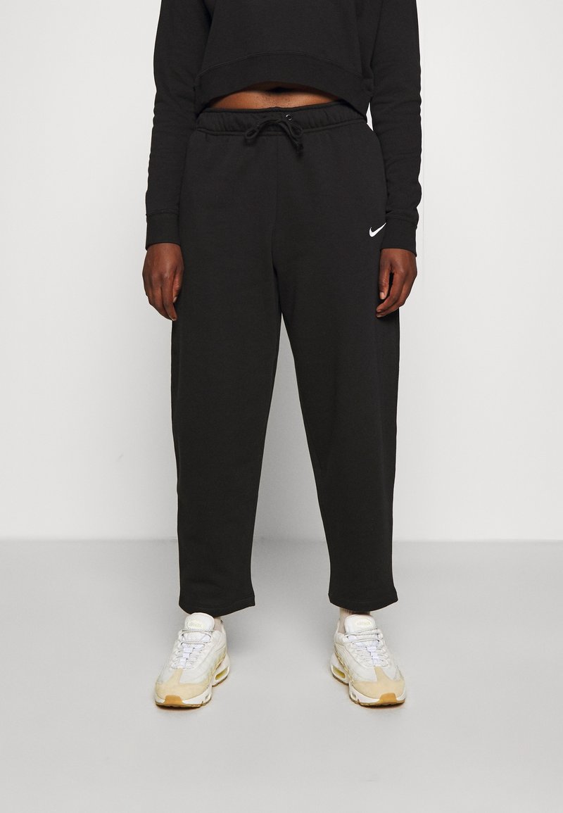 Svarta sweatpants med elastisk midja, sidofickor och en diskret vit Nike-logo på vänster lår, ihop med ljusa sneakers.