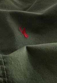 Tkanina w kolorze oliwkowym z teksturowaną powierzchnią, z czerwonym haftowanym logo jelenia w dolnej części.