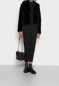 Veste en fausse fourrure noire avec col châle, associée à un pantalon noir ajusté et des bottines noires épaisses. Accessoires : un sac à main noir.
