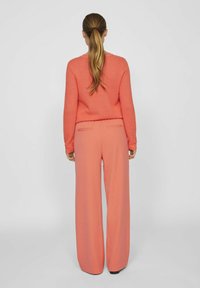 Žena s dlouhým copem, oblečená v korálovém pleteném svetru a širokých korálových kalhotách, stojící čelem k jednoduchému světle šedému pozadí.