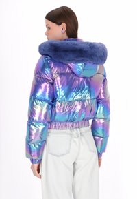 Perłowa kurtka puchowa z niebieskim kapturem z sztucznego futra, elastycznym dołem i obszernymi rękawami, w zestawie z jasnymi spodniami dżinsowymi.