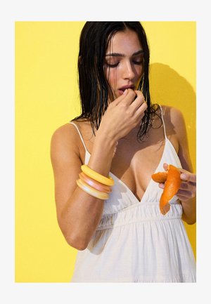 Femme aux cheveux mouillés portant une robe blanche et des bracelets colorés, épluchant et mangeant une orange sur un fond jaune vif.